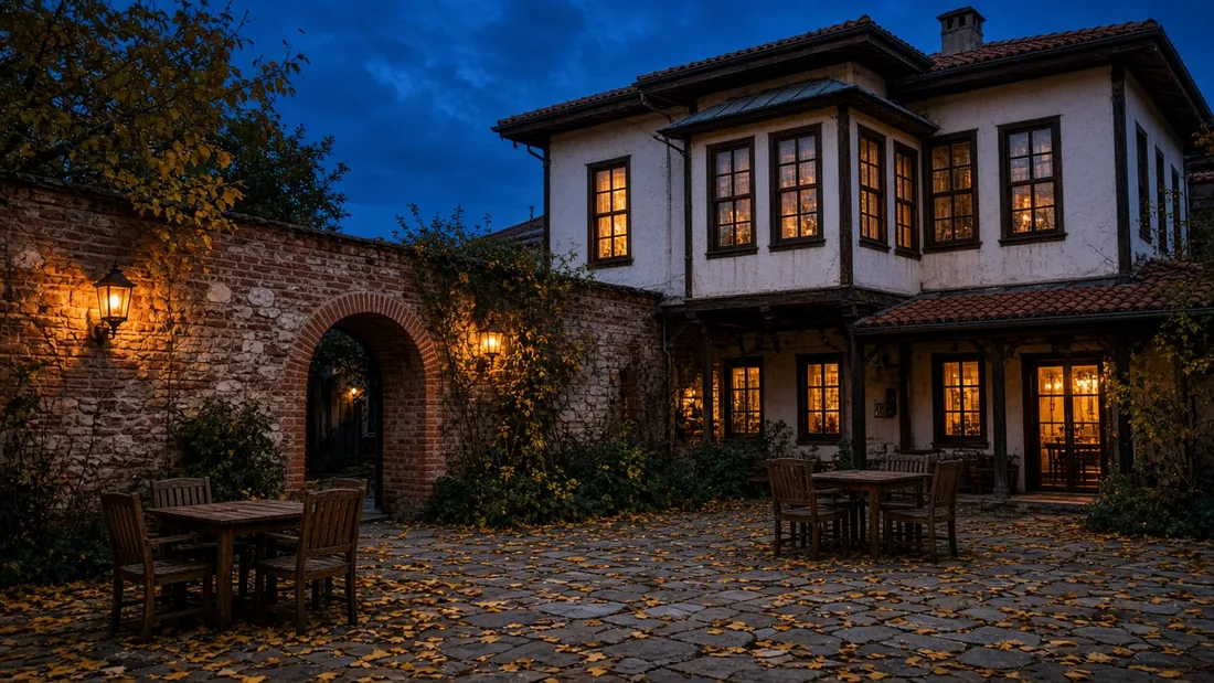 Akşam saatlerinde sıcak ışıklı fenerlerle aydınlanan taş bir avluda boş ahşap masalar ve yerde dökülmüş sarı yapraklar