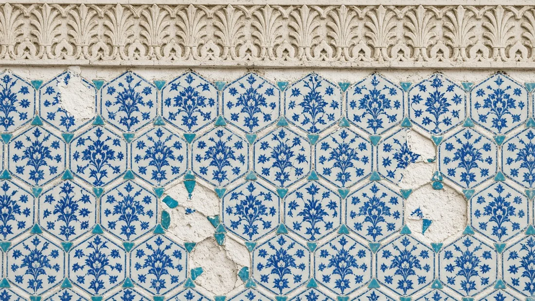 Beyaz zemin üzerine kobalt mavisi çiçek desenli altıgen çinilerin firuze üçgenlerle birleşerek oluşturduğu yıldızlı duvar yüzeyinin yakın planı, üstte kabartmalı palmet frizi